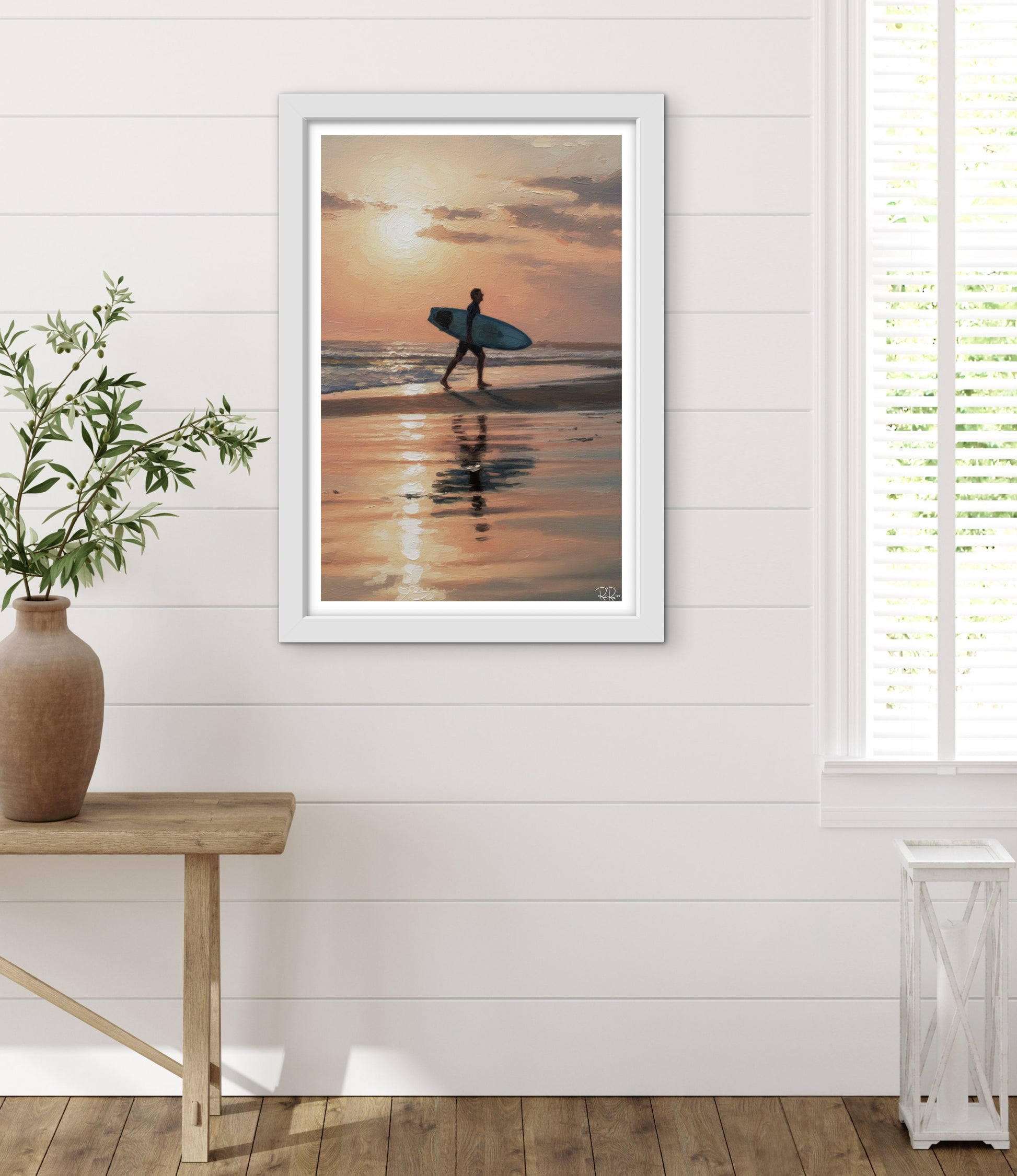 Framed artwork of a person with a surfboard walking on a beach at sunset, displayed in a room with a wooden table and plant.