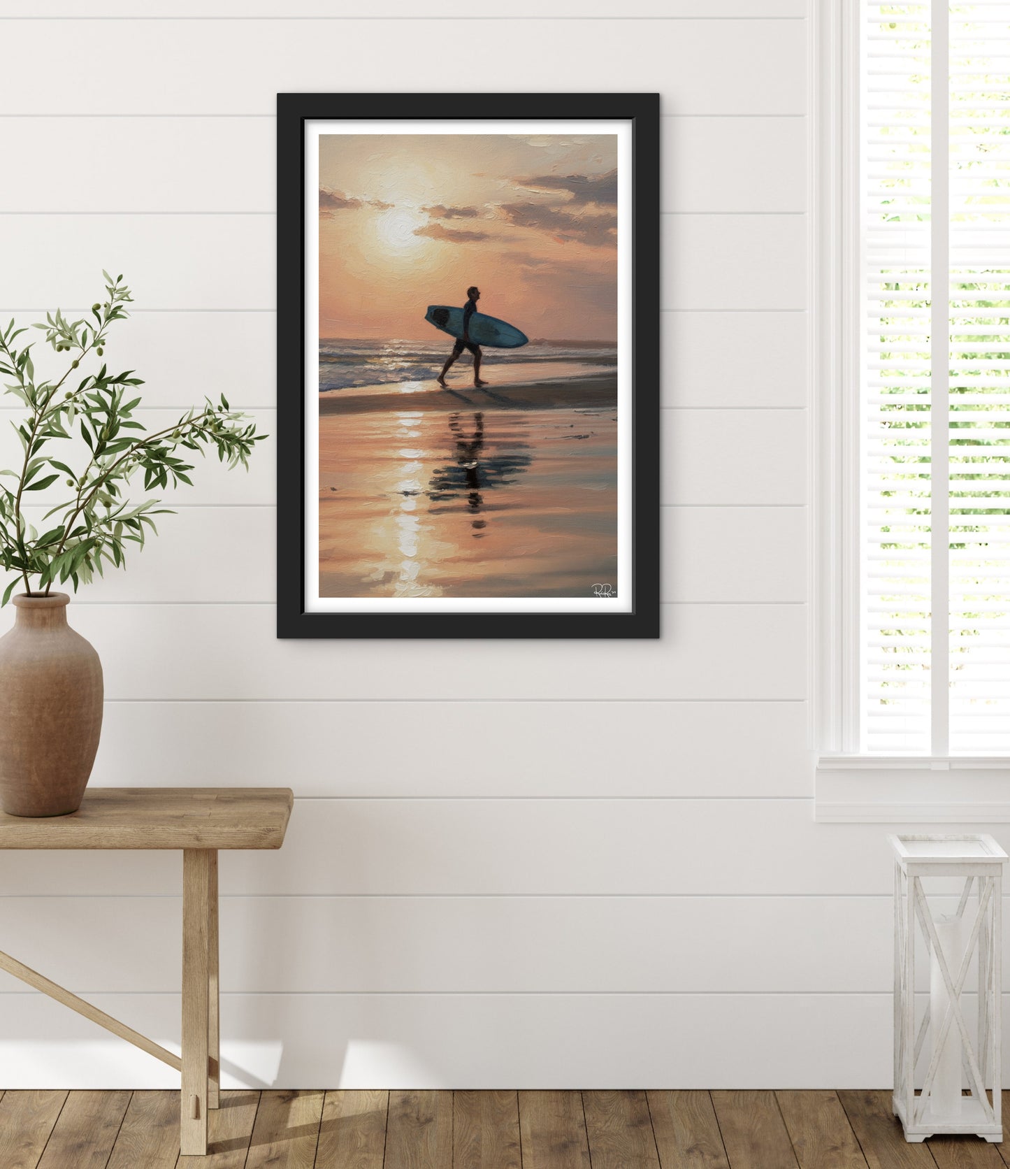 Framed artwork of a person with a surfboard at sunset on a beach, hanging on a white wall above a wooden table.