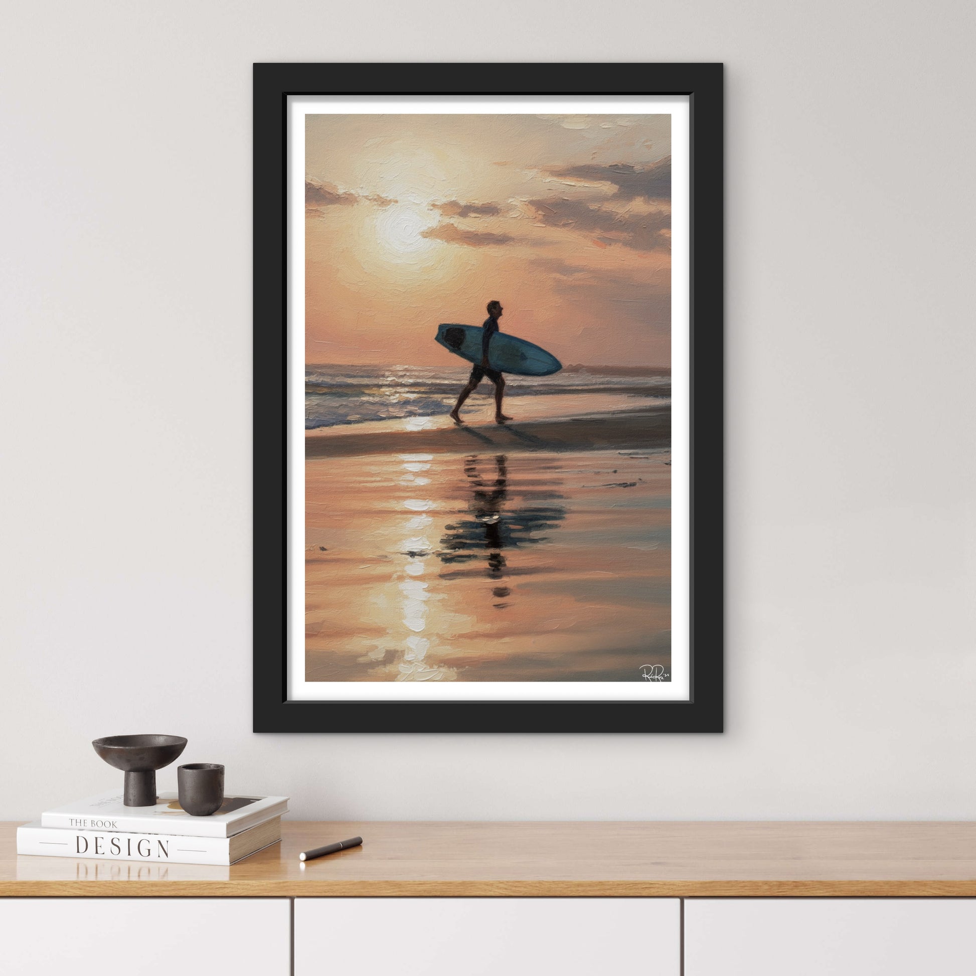 Framed artwork of a person with a surfboard walking on a beach at sunset, displayed on a wall above a wooden surface.