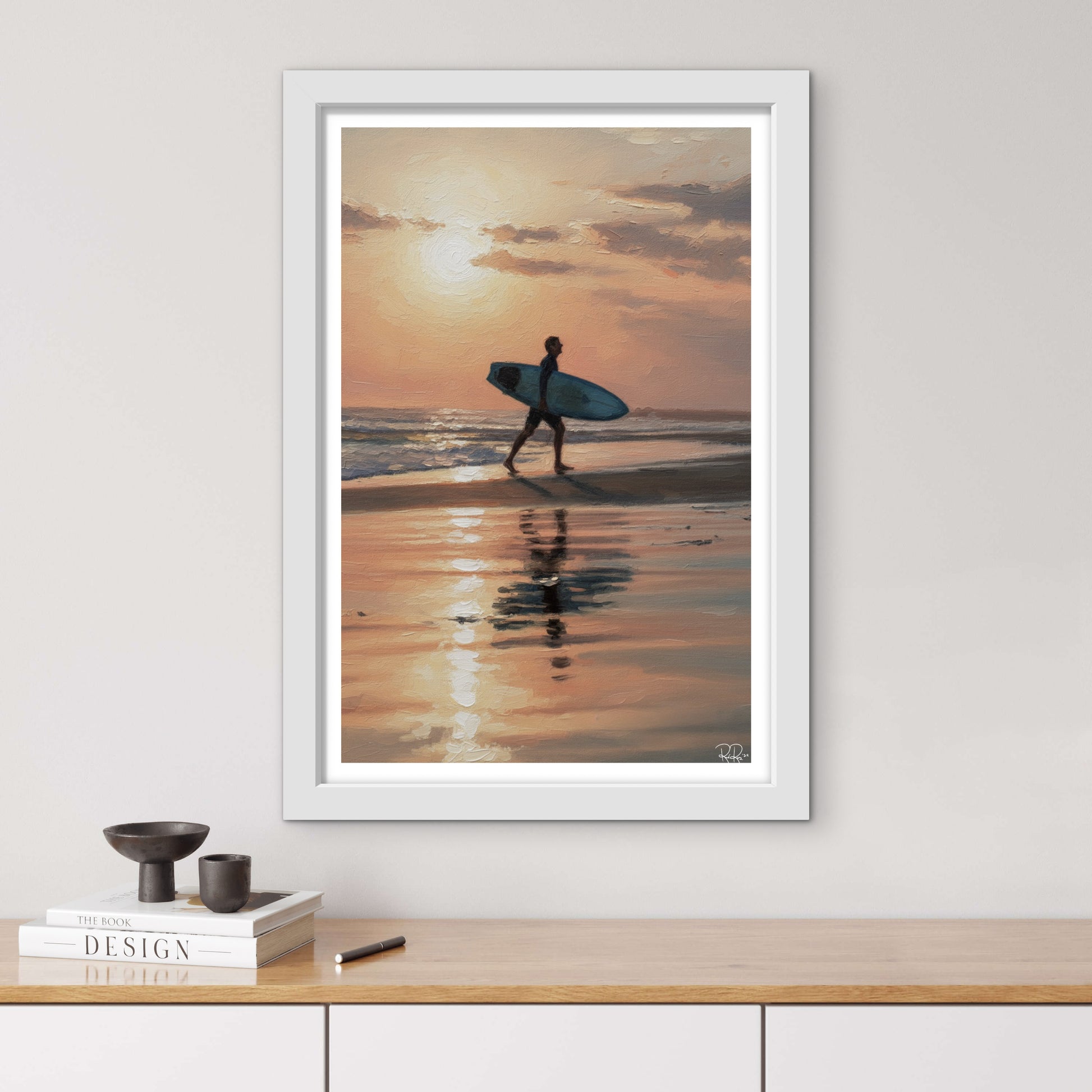 Framed artwork of a person with a surfboard walking on a beach at sunset, displayed on a wall above a wooden surface.