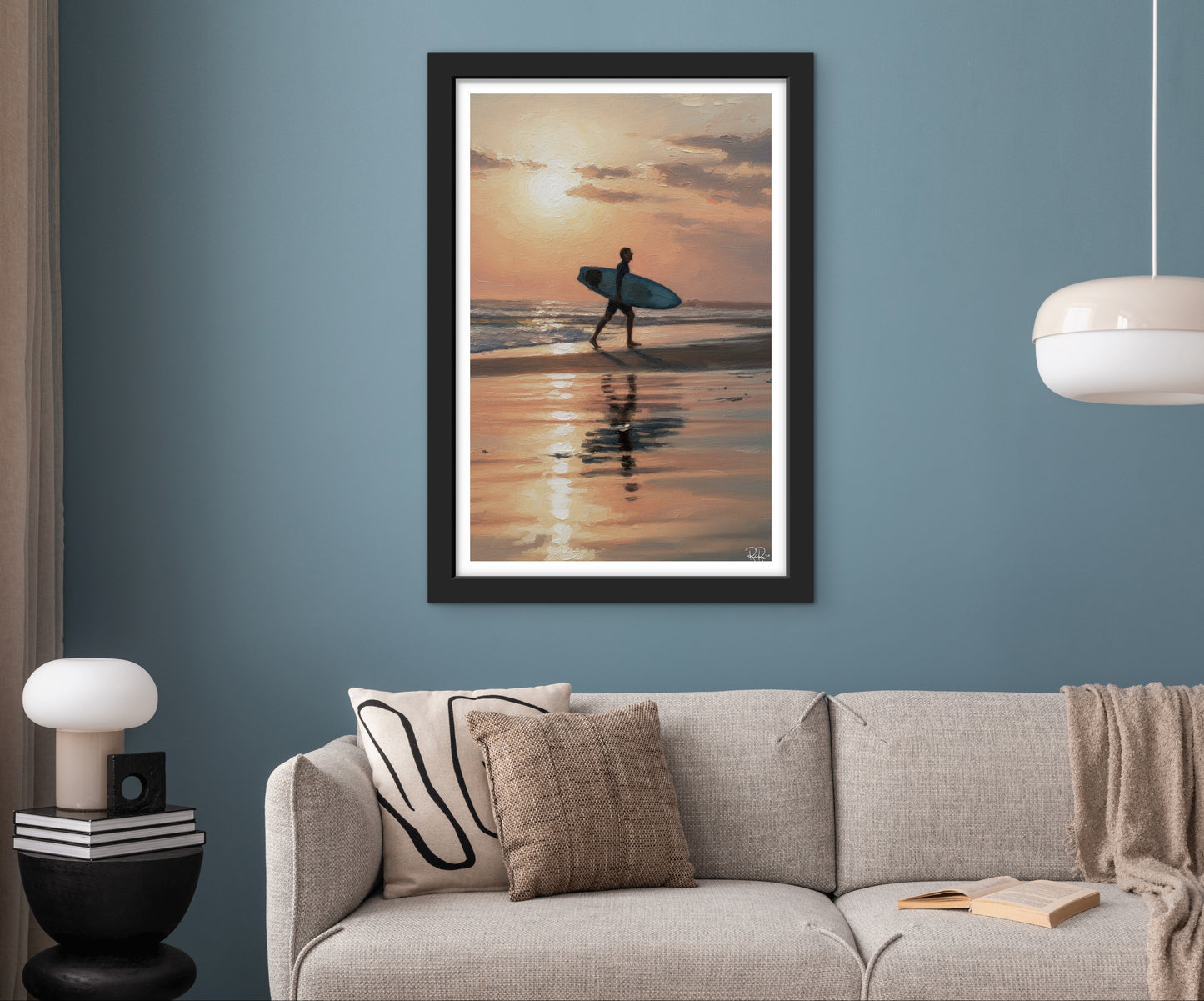 Framed artwork of a person with a surfboard on a beach at sunset above a sofa.
