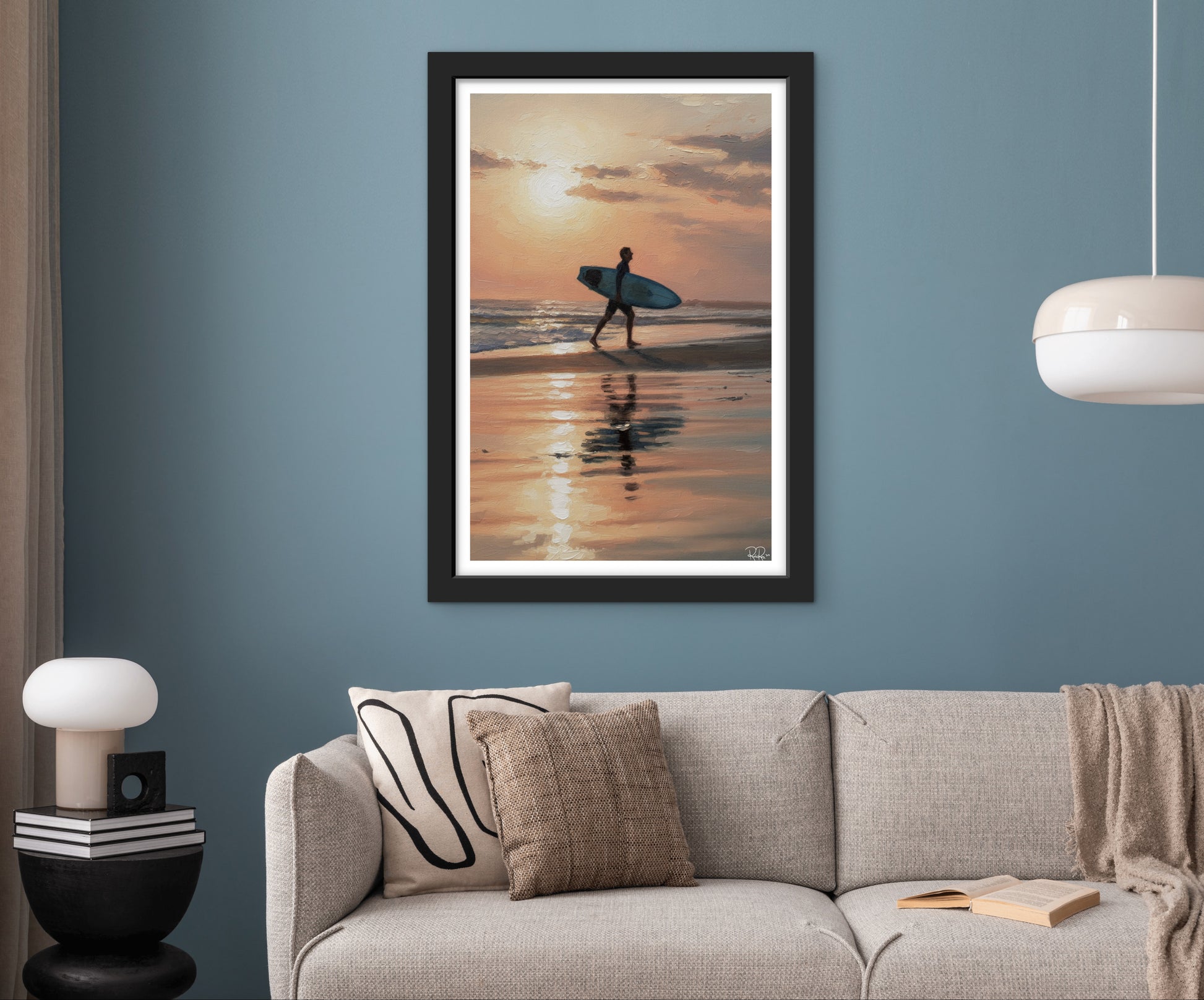 Framed artwork of a person with a surfboard on a beach at sunset above a sofa.