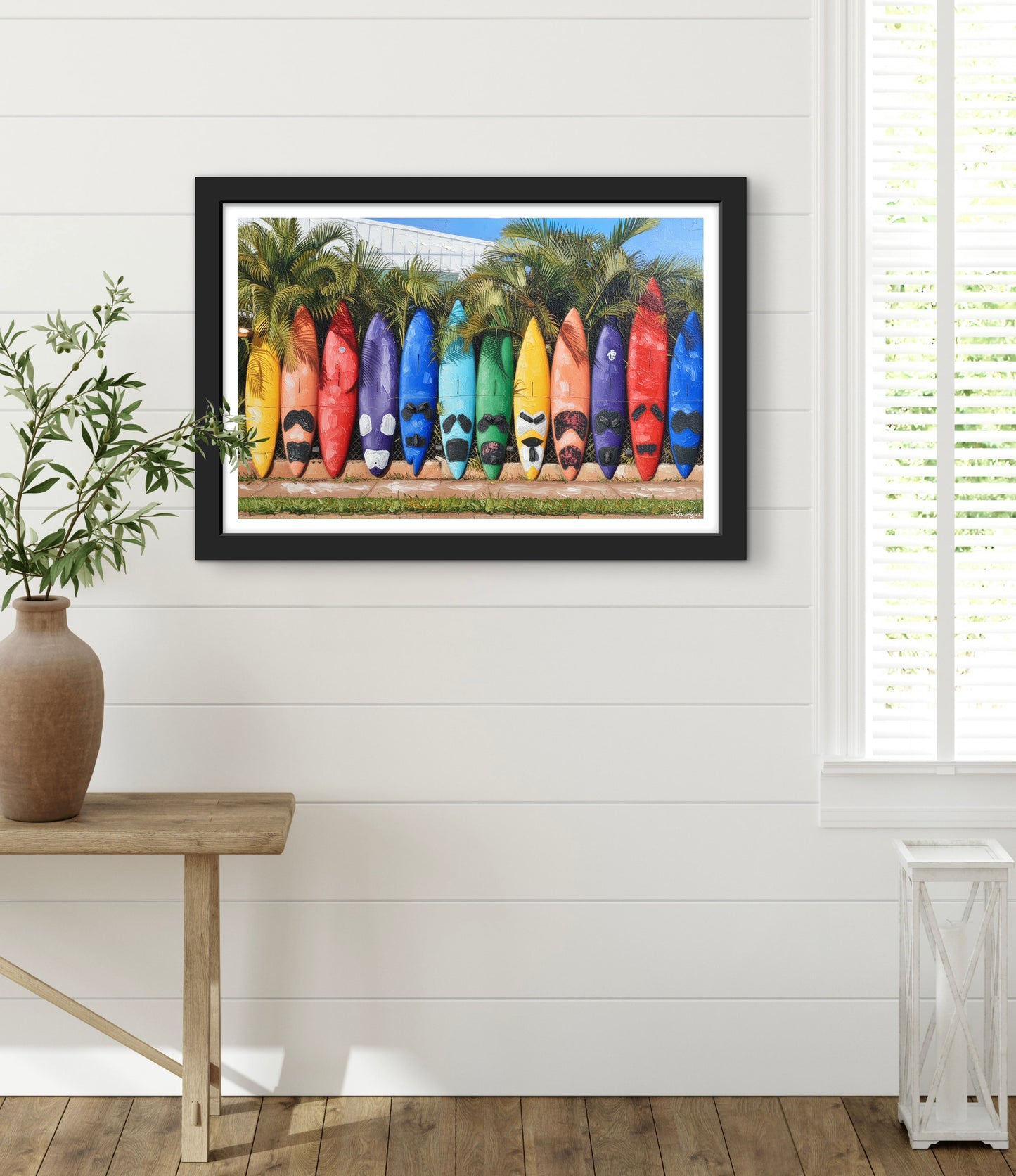 Framed artwork of colorful surfboards on a white wall with a wooden table and plant in the foreground.