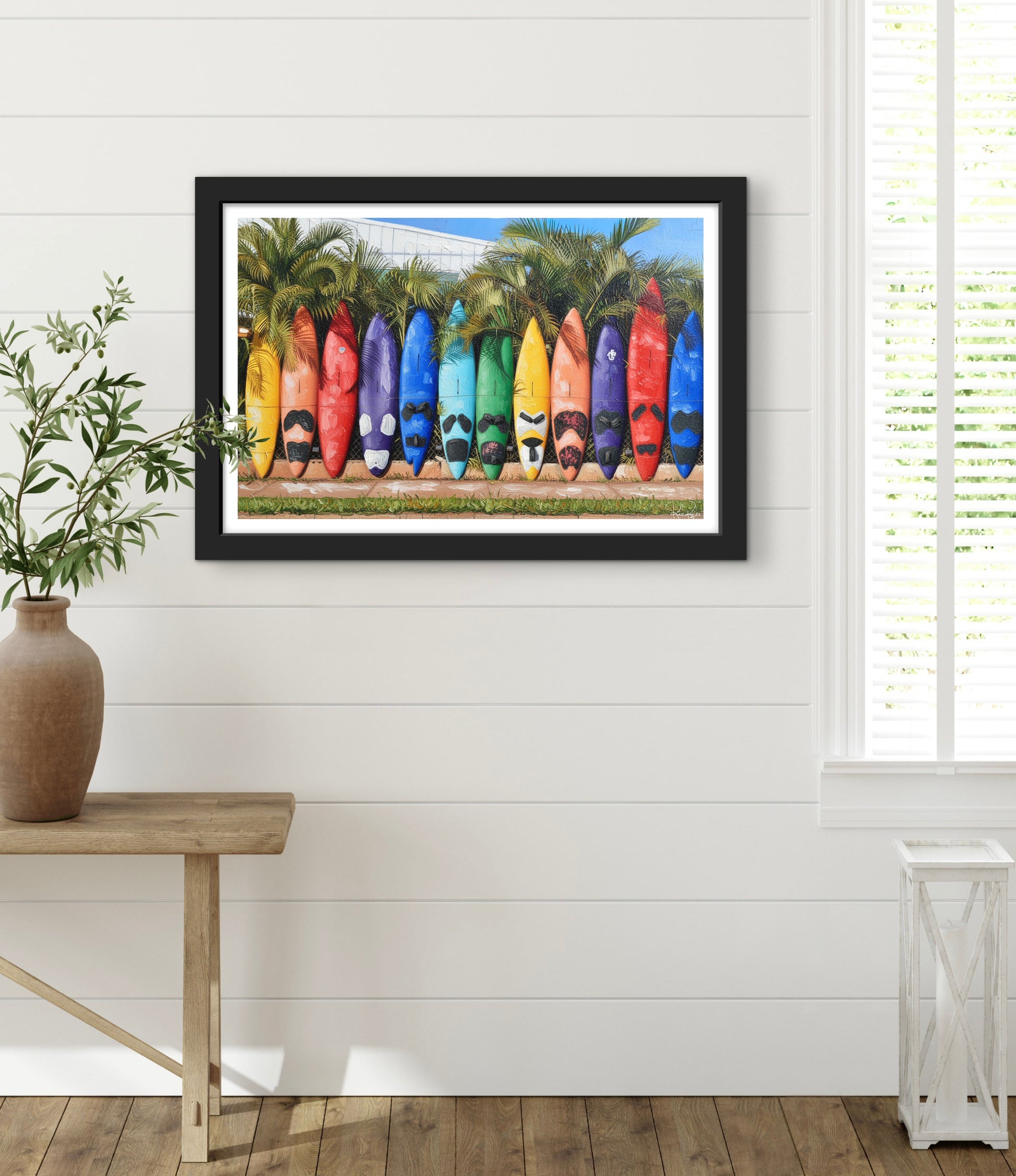 Framed artwork of colorful surfboards on a white wall with a wooden table and plant in the foreground.