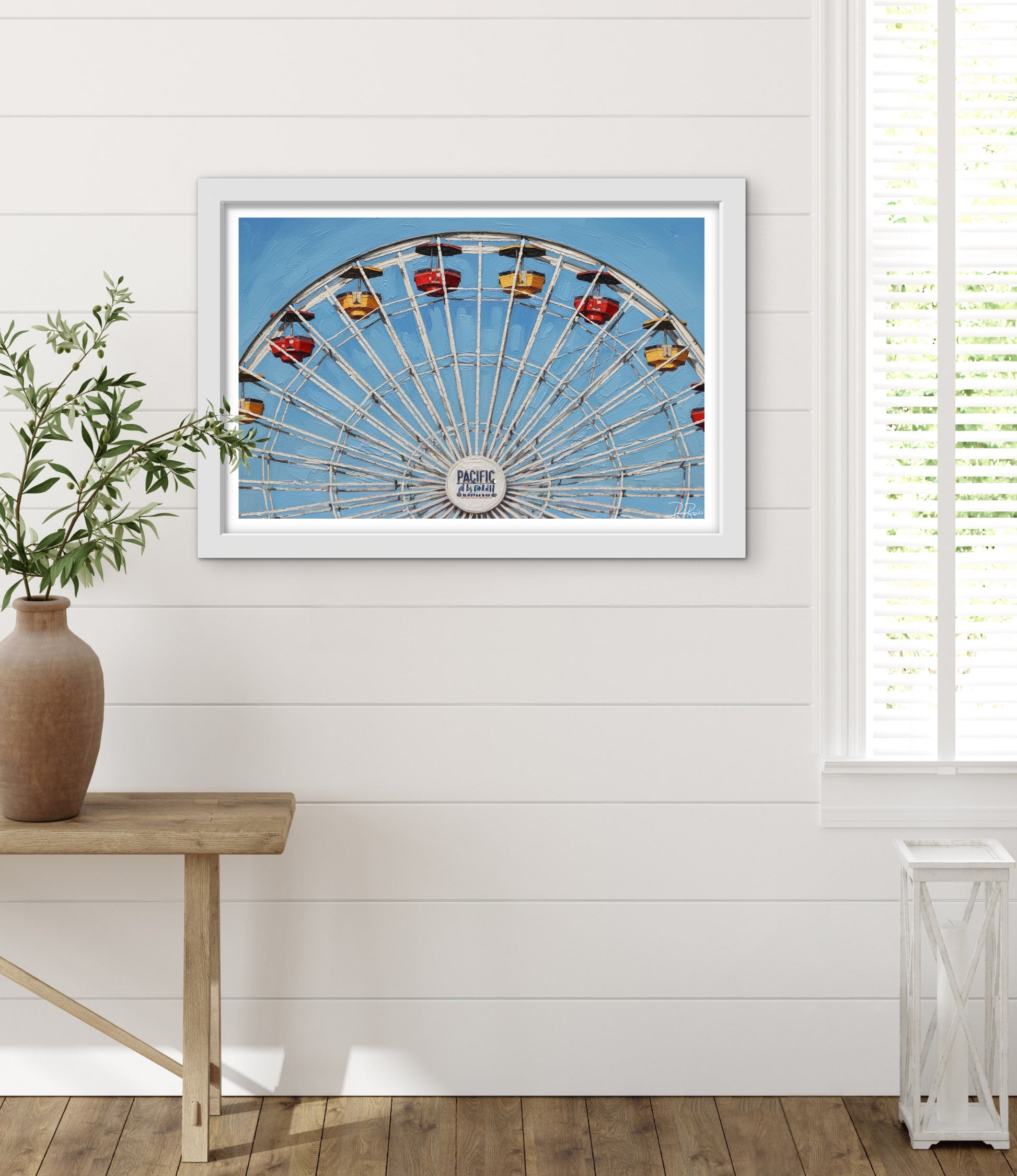 Framed picture of a Ferris wheel on a white wall with a plant and wooden table in the foreground.